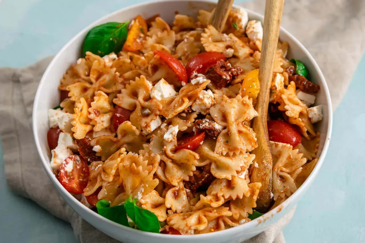 Farfalle-Nudelsalat mit frischen Tomaten, Mozzarella und sonnengetrockneten Tomaten, garniert mit Basilikum.