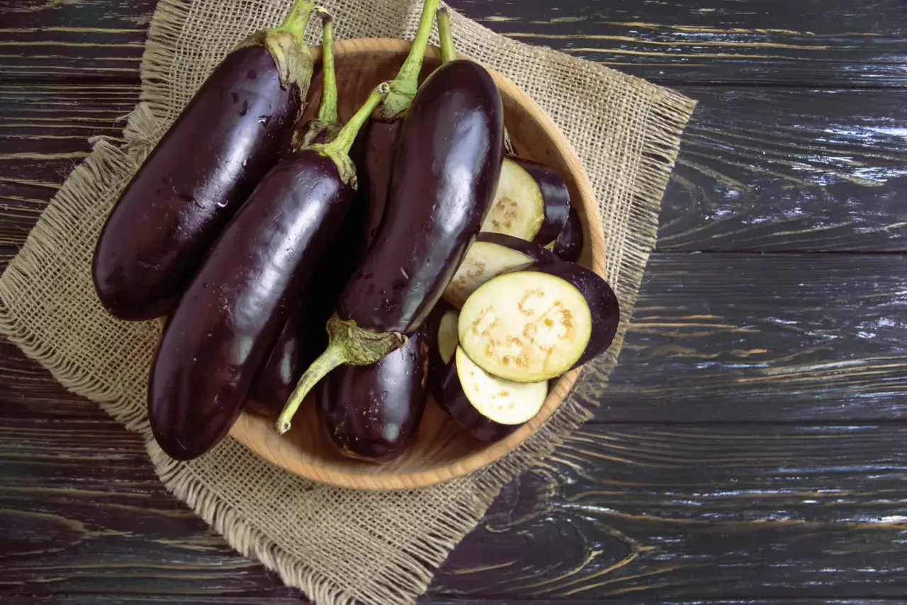 Einige ganze Auberginen liegen neben geschnittenen Scheiben in einer Holzschale. Man sieht das helle Fruchtfleisch der Aubergine innen.