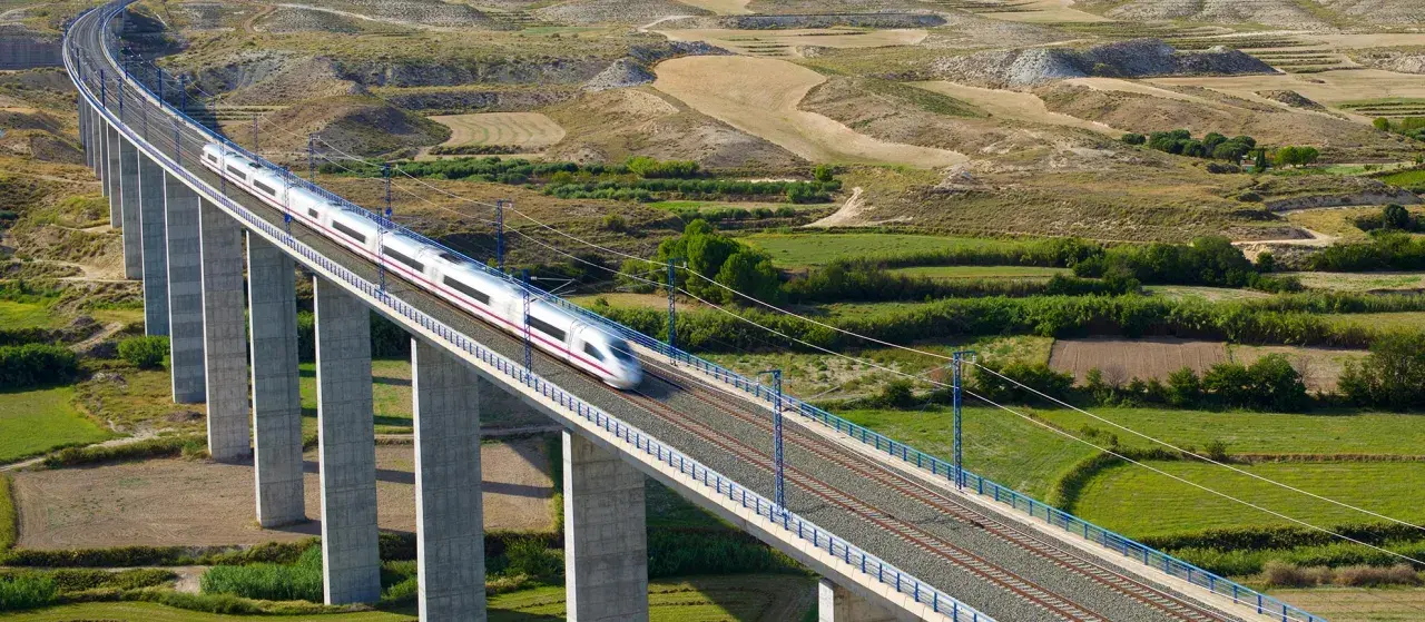 Un tren de alta velocidad cruza un viaducto sobre un paisaje rural. El tren, que parece ir de Lleida a Madrid, es plateado con una franja morada.