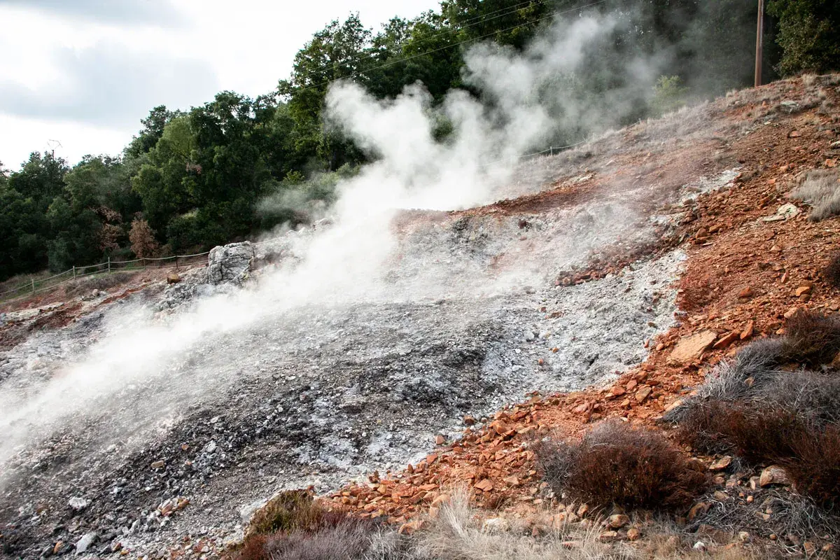 Fumarole di Monterotondo Marittimo emettono vapore su un pendio roccioso e arido, con vegetazione secca e alberi sullo sfondo.