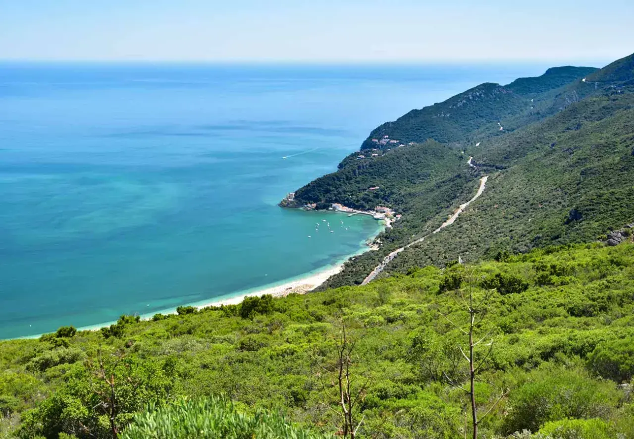 Costa verde en el sur de Lisboa, con mar turquesa, playa, barcos y montañas cubiertas de vegetación.