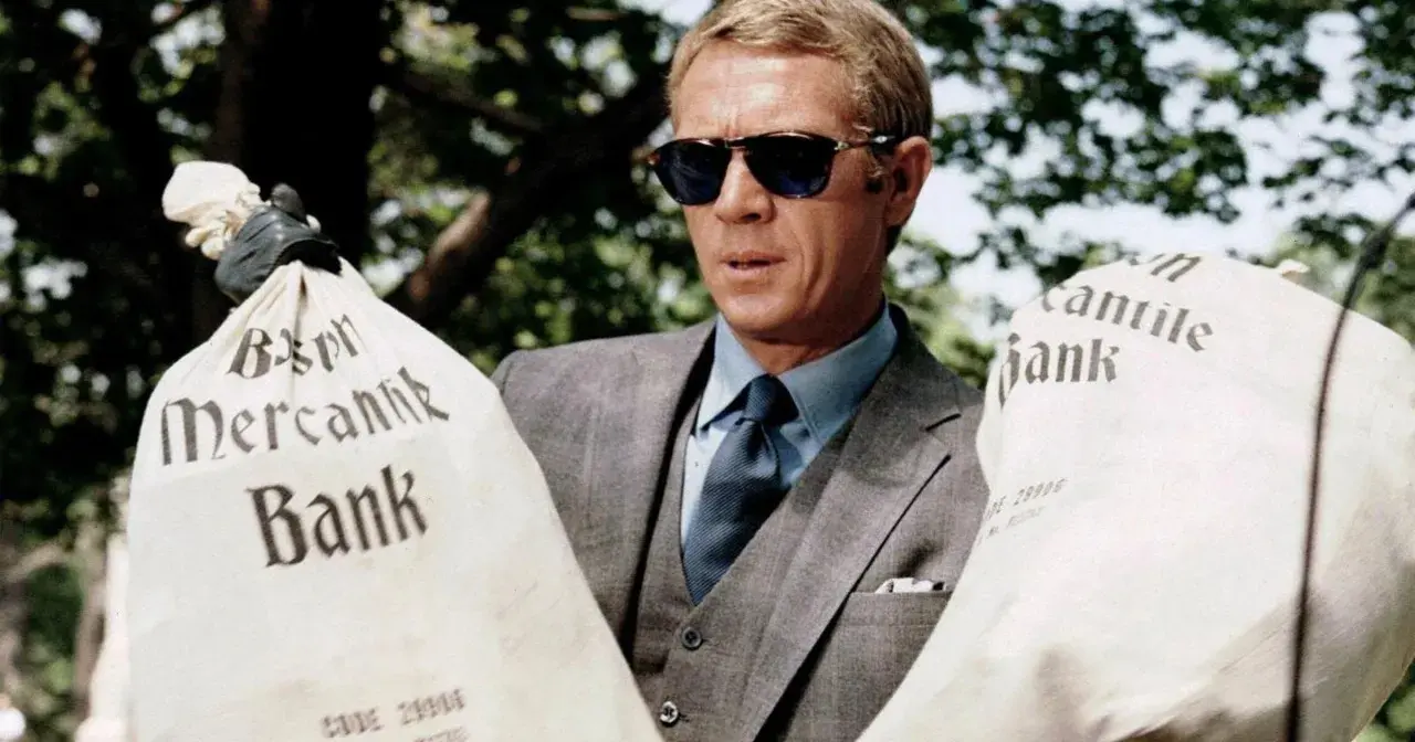 Steve McQueen w okularach przeciwsłonecznych, w eleganckim garniturze, trzyma worki z napisem "Mercantile Bank". Scena z filmu "Afera Thomasa Crowna".