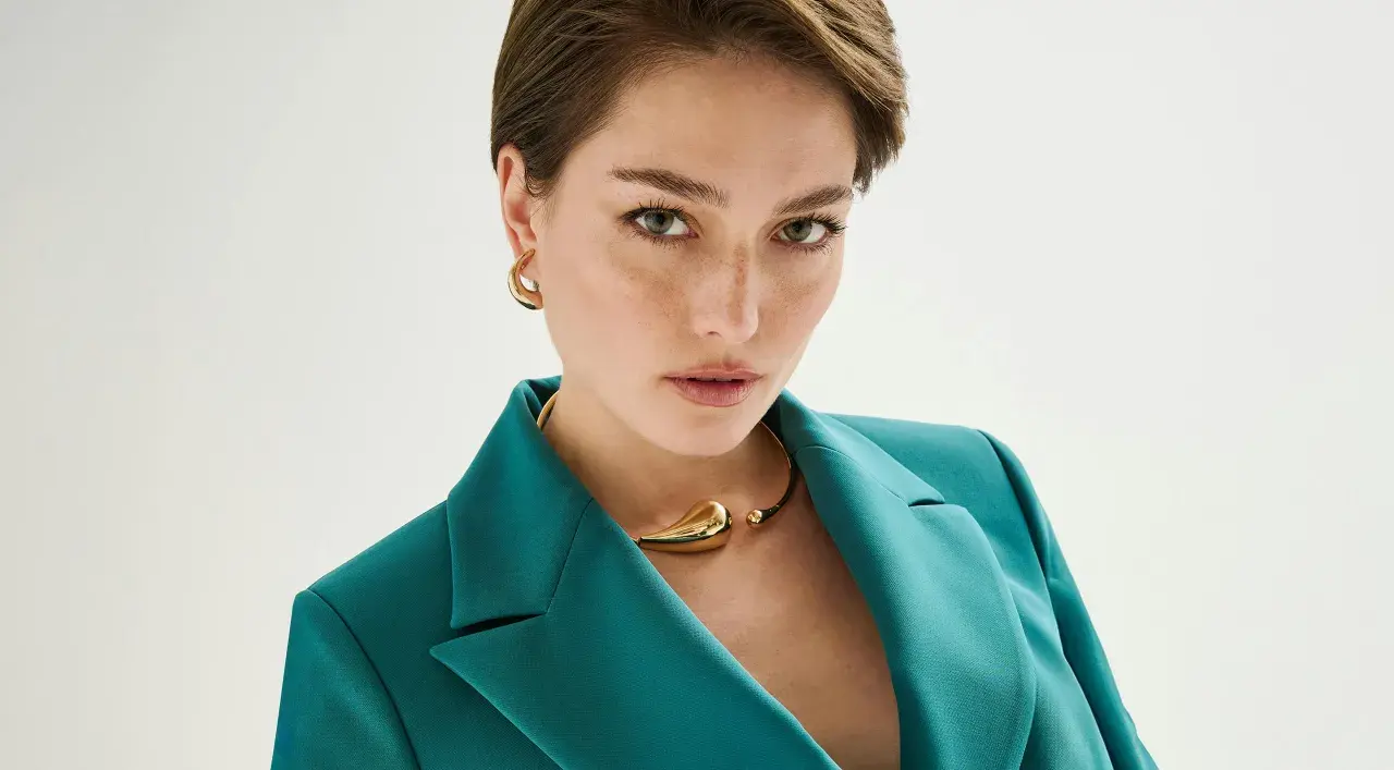Mujer con un elegante corte pixie, viste un blazer verde azulado y un collar dorado.