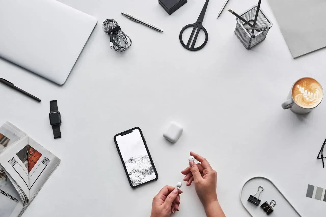 Dłonie trzymające białe słuchawki AirPods obok smartfona i etui.