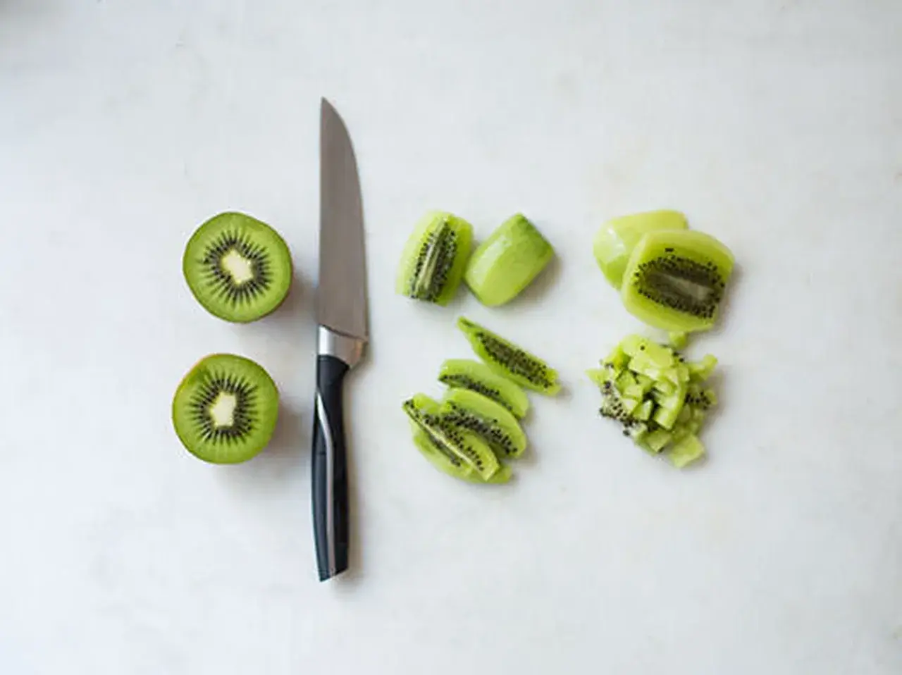 Ein Messer liegt neben zwei halbierten Kiwis. Daneben sind die Früchte in Scheiben und Würfel geschnitten, bereit zum kiwi schneiden.