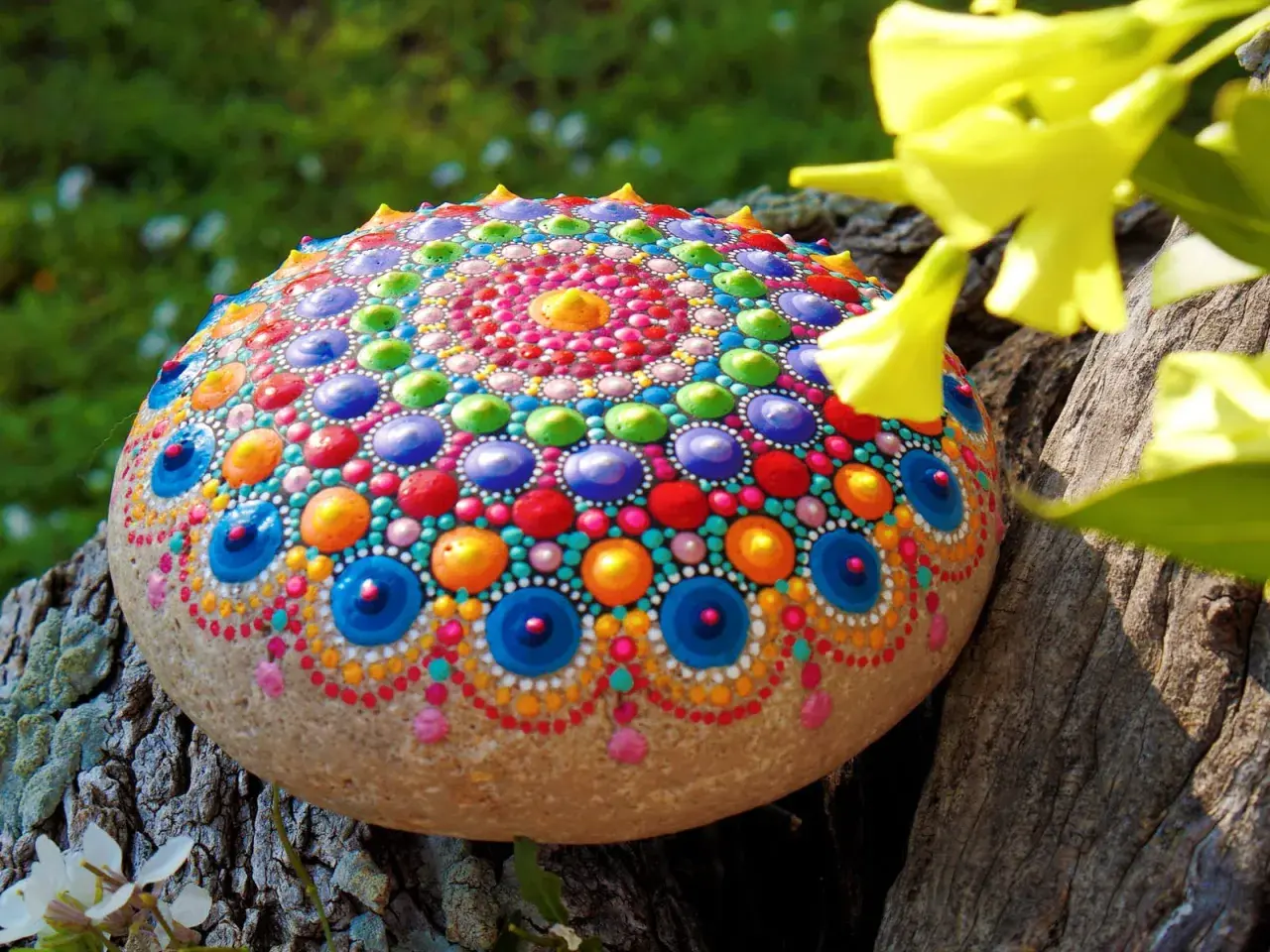 Bunte bemalte Steine mit Mandala-Muster, die auf einem Baumstamm liegen, umgeben von grünem Gras und gelben Blüten.