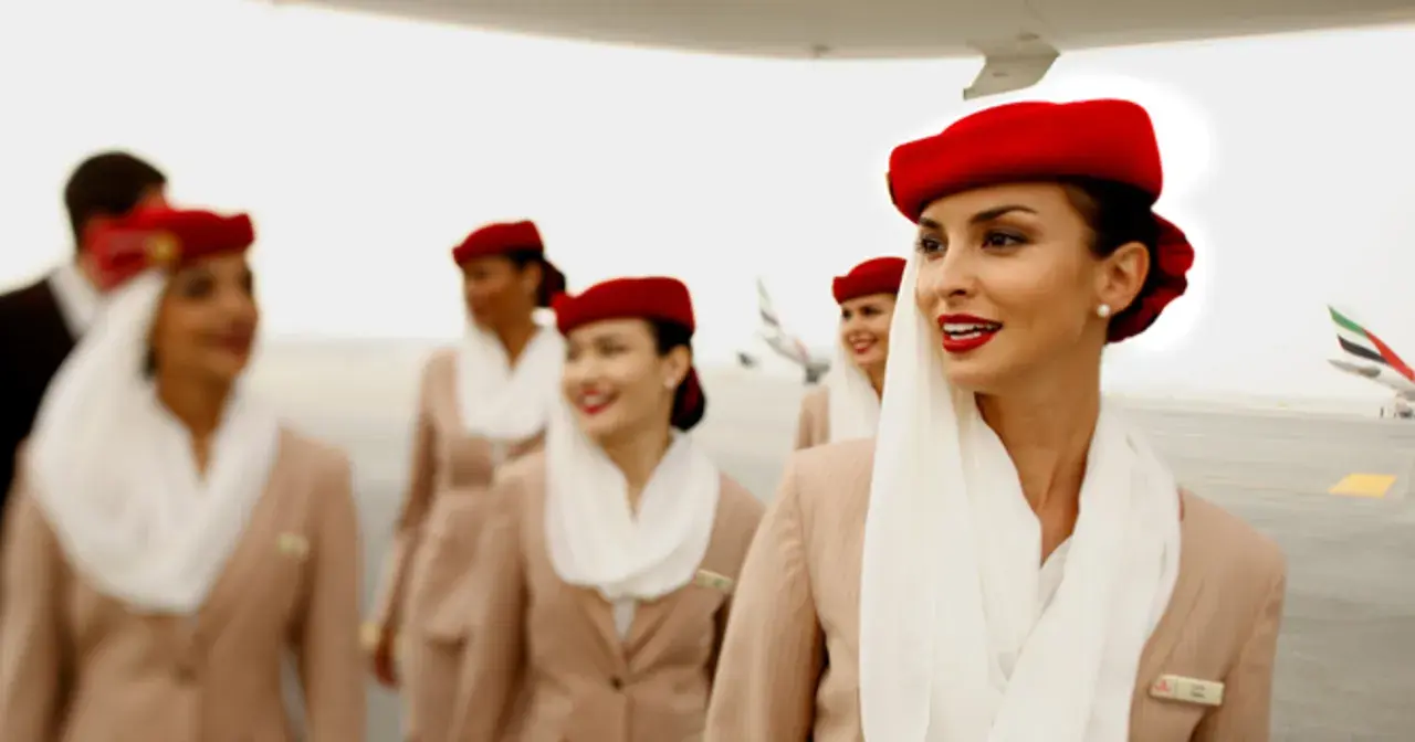 Uśmiechnięte stewardessy Emirates w czerwonych beretach i beżowych uniformach idą po płycie lotniska.