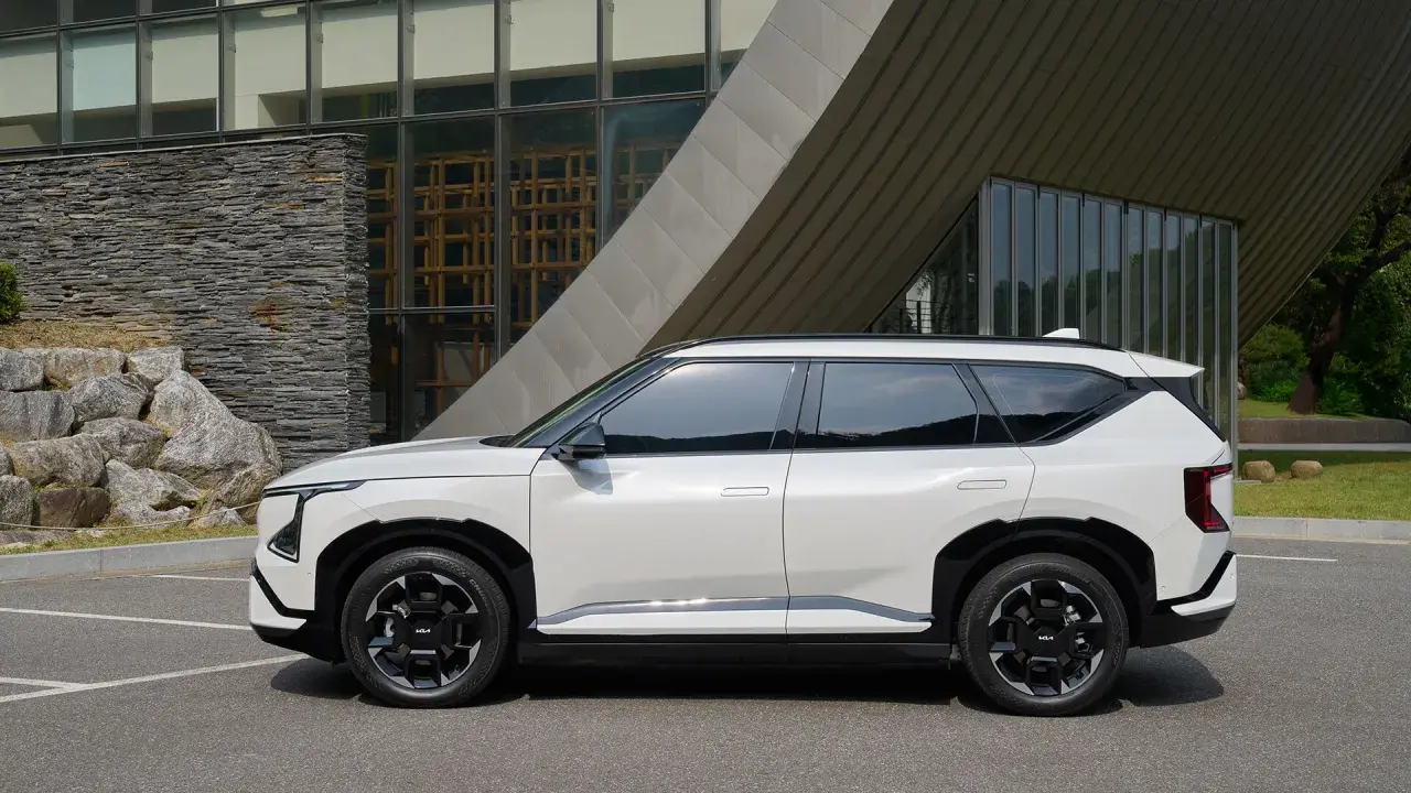 Nowy, biały suv Kia EV9 stoi zaparkowany przed nowoczesnym budynkiem z kamienną fasadą i dużymi oknami.
