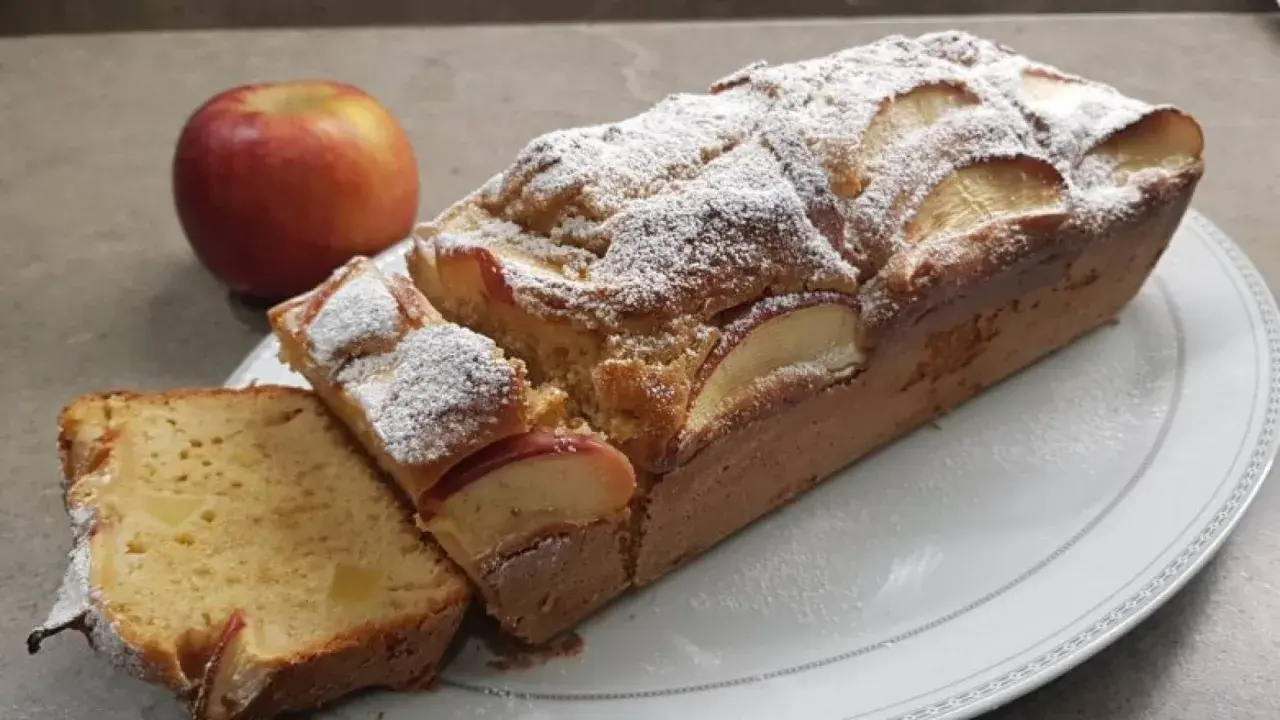 Un delizioso plumcake alle mele, cosparso di zucchero a velo, con una mela rossa sullo sfondo.