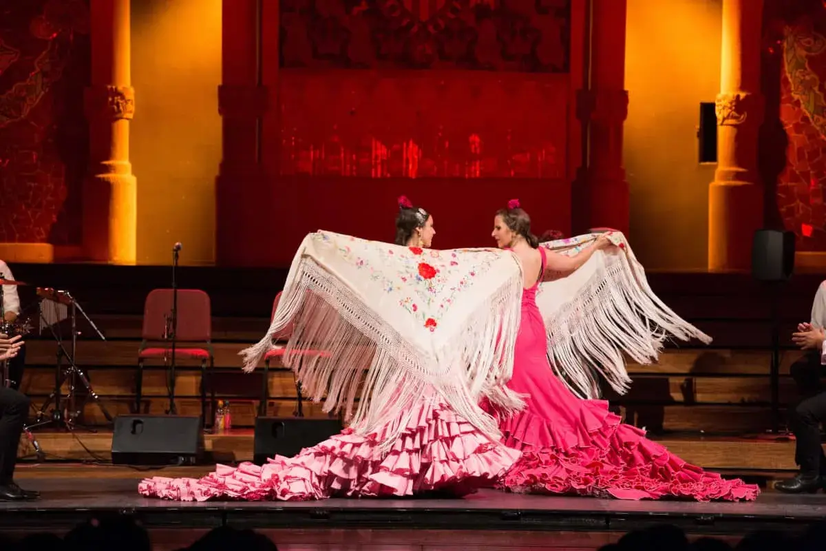 Dos bailarinas de flamenco, con vestidos rosas y mantones bordados, despliegan su arte. El escenario tiene un aire de tener aje.