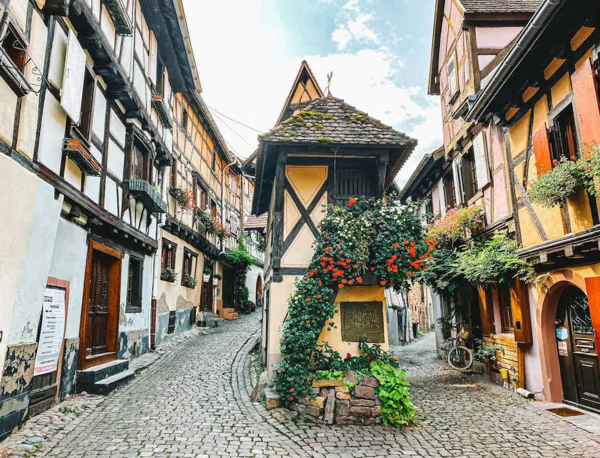 Kopfsteinpflastergasse im Elsass mit Fachwerkhäusern, Blumen und einem Fahrrad. Ein perfekter Ort für Geheimtipps im Elsass.