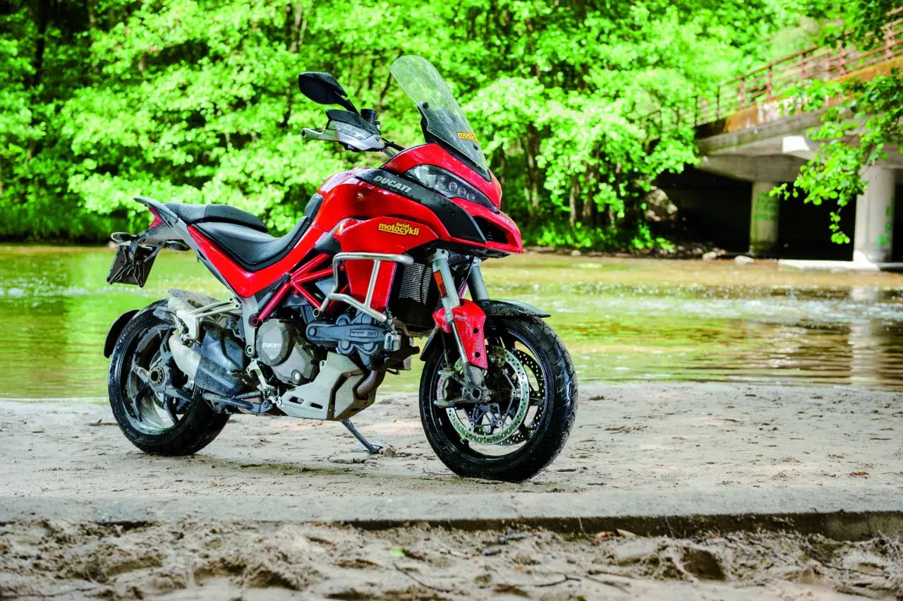 Czerwony Ducati Multistrada 1200S zaparkowany na piaszczystym brzegu rzeki, otoczony bujną zielenią.