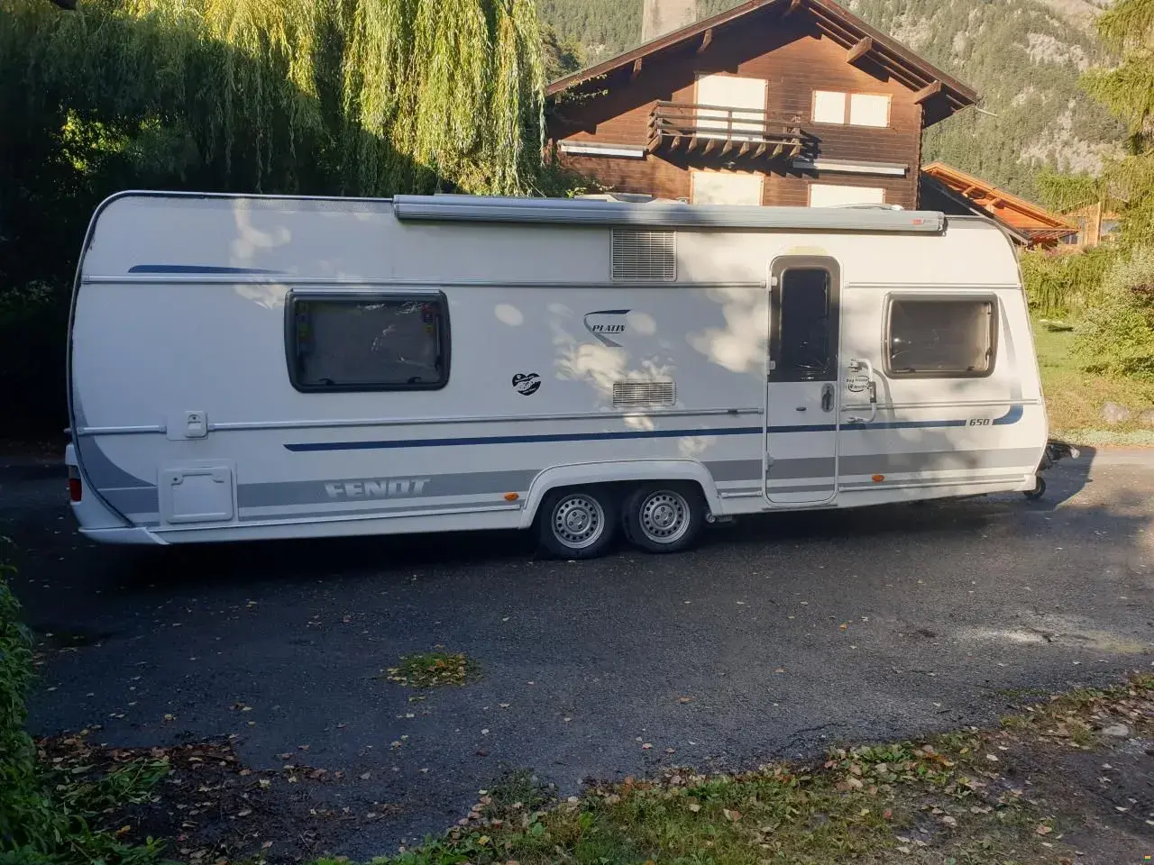 Caravane Fendt blanche garée devant une maison en bois, avec des arbres en arrière-plan.