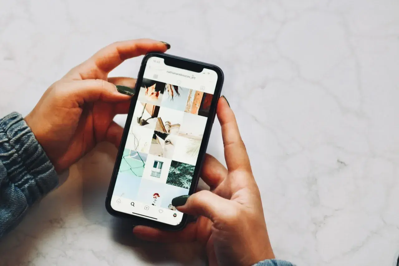 Dłonie trzymające telefon, na ekranie widoczny profil Instagrama z moimi pracami. Niestety, moje urządzenie iPhone niedostępne.