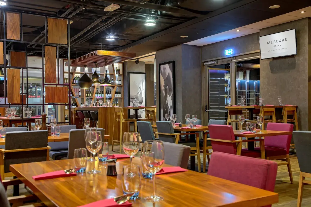 Elegancka restauracja w hotelu Mercure Wrocław Centrum. Drewniane stoły, czerwone i szare krzesła, półki z winami.