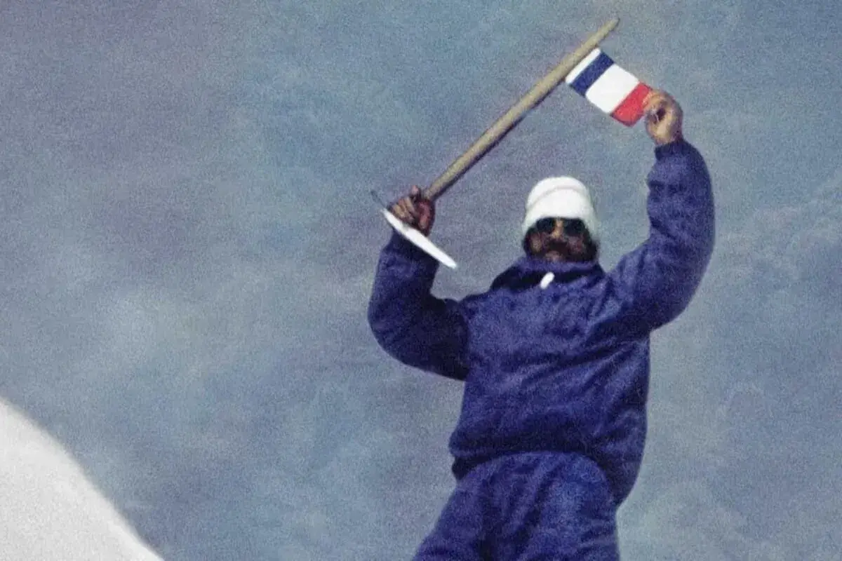 Les mains de Maurice Herzog brandissent un drapeau français au sommet d'une montagne enneigée, symbole d'une victoire alpine.
