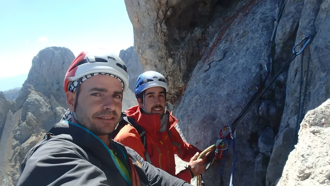 Dos escaladores sonríen en la cara del Naranjo de Bulnes, equipados con cascos y arneses, listos para la aventura.
