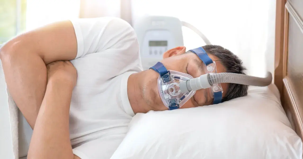 Mężczyzna śpi z maską CPAP, która pomaga w leczeniu bezdechu sennego. Urządzenie zapewnia ciągłe dodatnie ciśnienie w drogach oddechowych, łagodząc objawy.