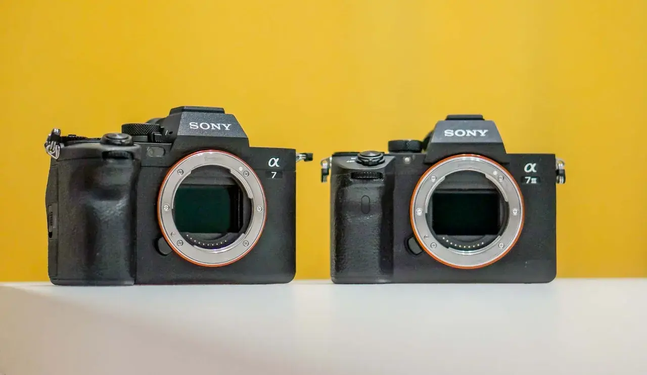 Dwa aparaty Sony Alpha, jeden z nich to sony a7 iiis, stoją obok siebie na białej powierzchni, na żółtym tle.