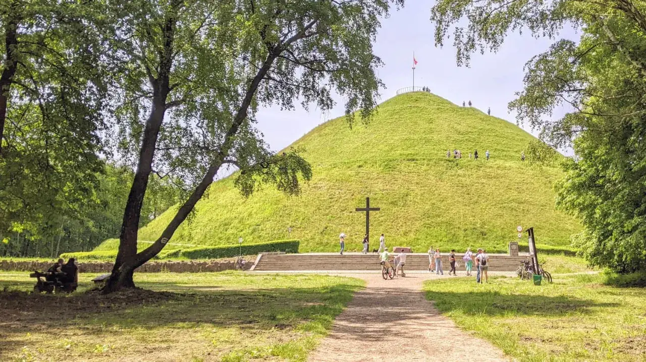 Zielony Kopiec Piłsudskiego z krzyżem i ludźmi spacerującymi po zboczu.