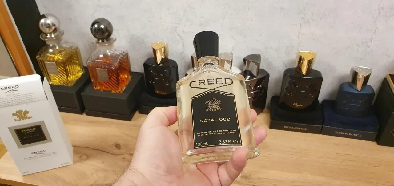Kolekcja perfum Creed, w tym Creed Royal Oud, prezentowana na drewnianym blacie. W dłoni trzymana jest butelka Creed Royal Oud.