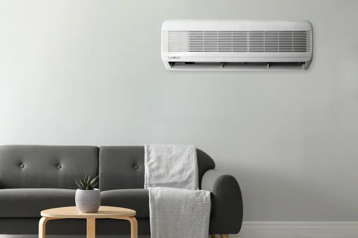 Unidad de aire acondicionado montada en la pared, cumpliendo la normativa aire acondicionado. Sofá gris con mantas y mesa auxiliar con planta.