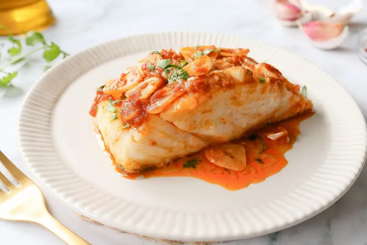 Delicioso bacalao a la llauna con salsa de tomate y ajo, servido en un plato blanco con un tenedor dorado.
