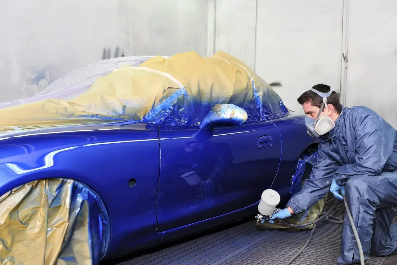 Un carrozziere con maschera spruzza vernice blu su un'auto coperta. Consulta il listino prezzi verniciatura auto per i costi.