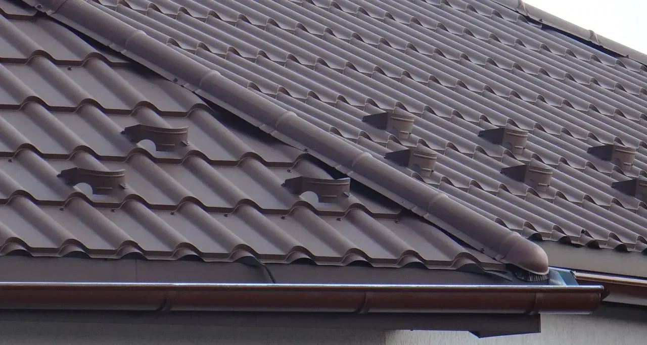 Brązowy dach z dachówki ceramicznej, widoczne elementy systemu rynnowego i wentylacyjnego. Jak zrobić zakończenie gąsiora?