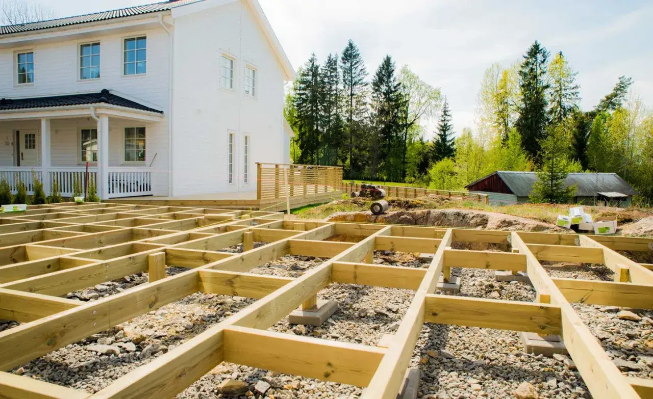 Grundarbete för att bygga altan på mark. Träkonstruktionen är på plats framför ett vitt hus.