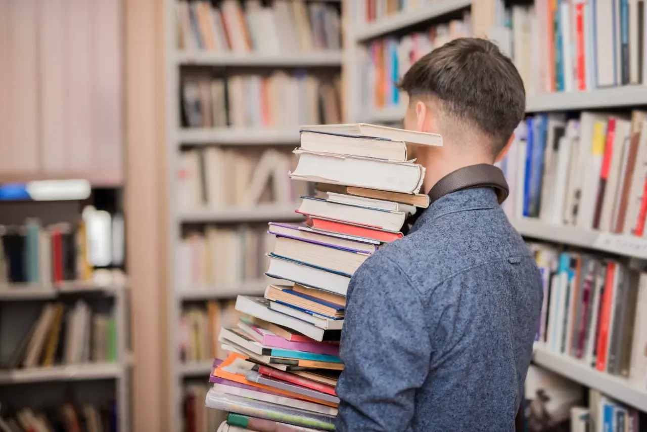 Młody mężczyzna w bibliotece dźwiga stos książek, symbolizując przejście od statusu ucznia do statusu studenta.