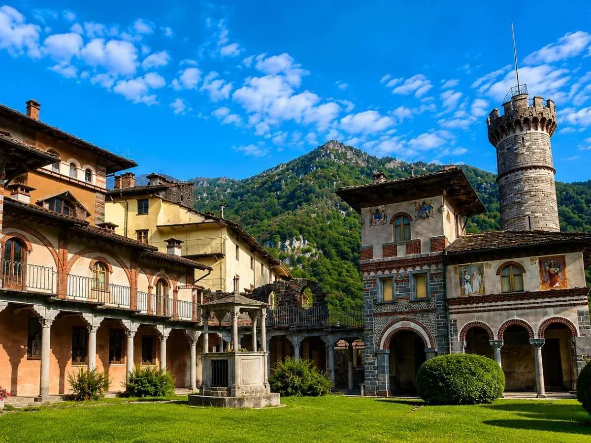 Cortile di un complesso architettonico con torre, affreschi e un pozzo, immerso nel verde. Sembra un luogo avvolto da un'aura di mistero, quasi una rosazza esoterica.