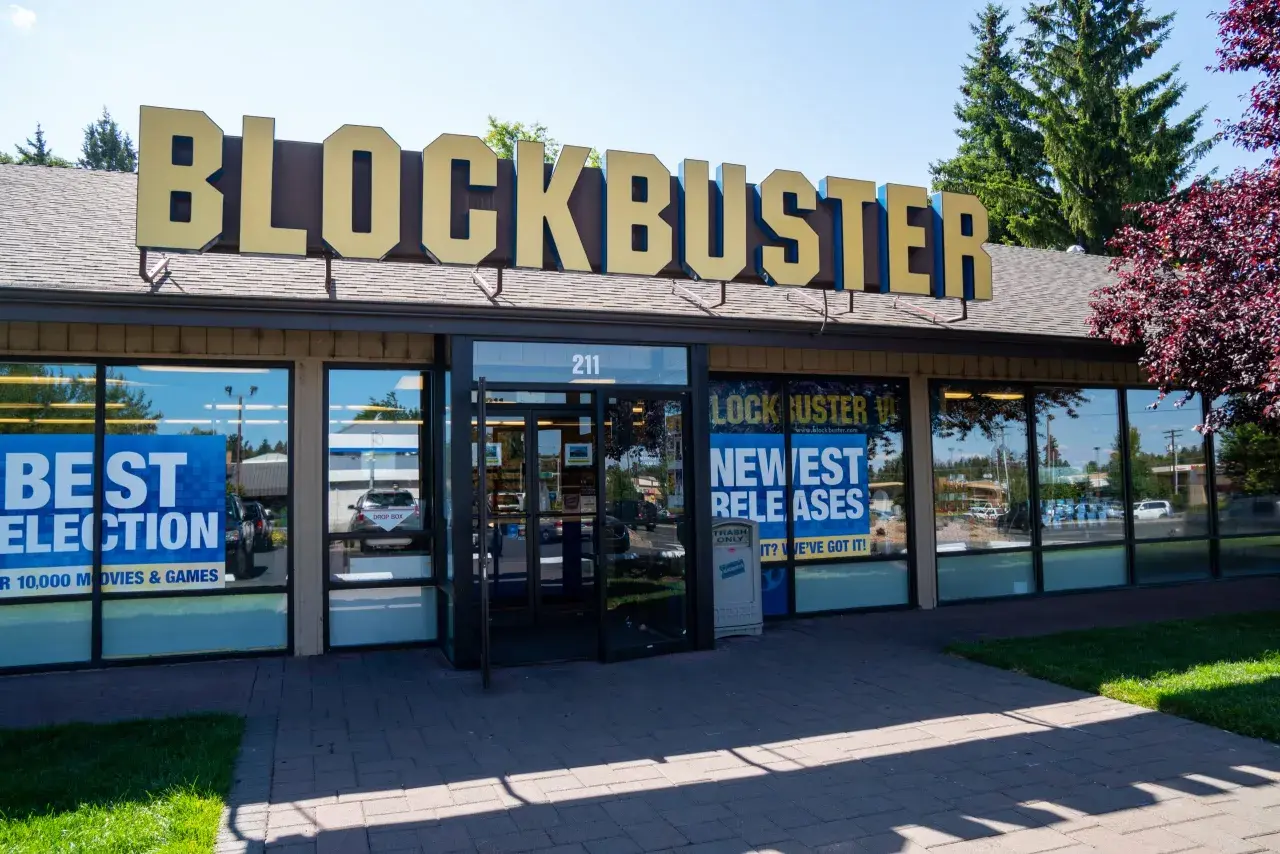 L'insegna dorata del negozio Blockbuster domina l'edificio. Vetrine mostrano "Best Selection" e "Newest Releases", un richiamo nostalgico ai tempi d'oro del blockbuster film.