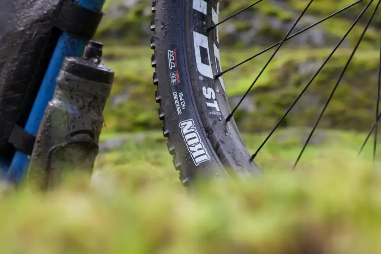 Close-up of a muddy bike tire, showing the "Maxxis Ikon" branding. This tire is ready for a challenging trail review.