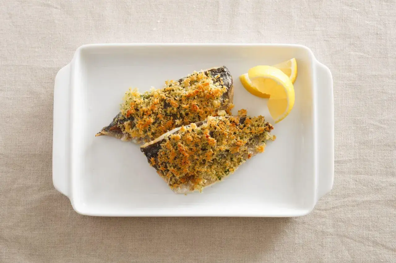 Two fillets of aji fish, baked with a golden breadcrumb topping, served with lemon wedges on a white dish.