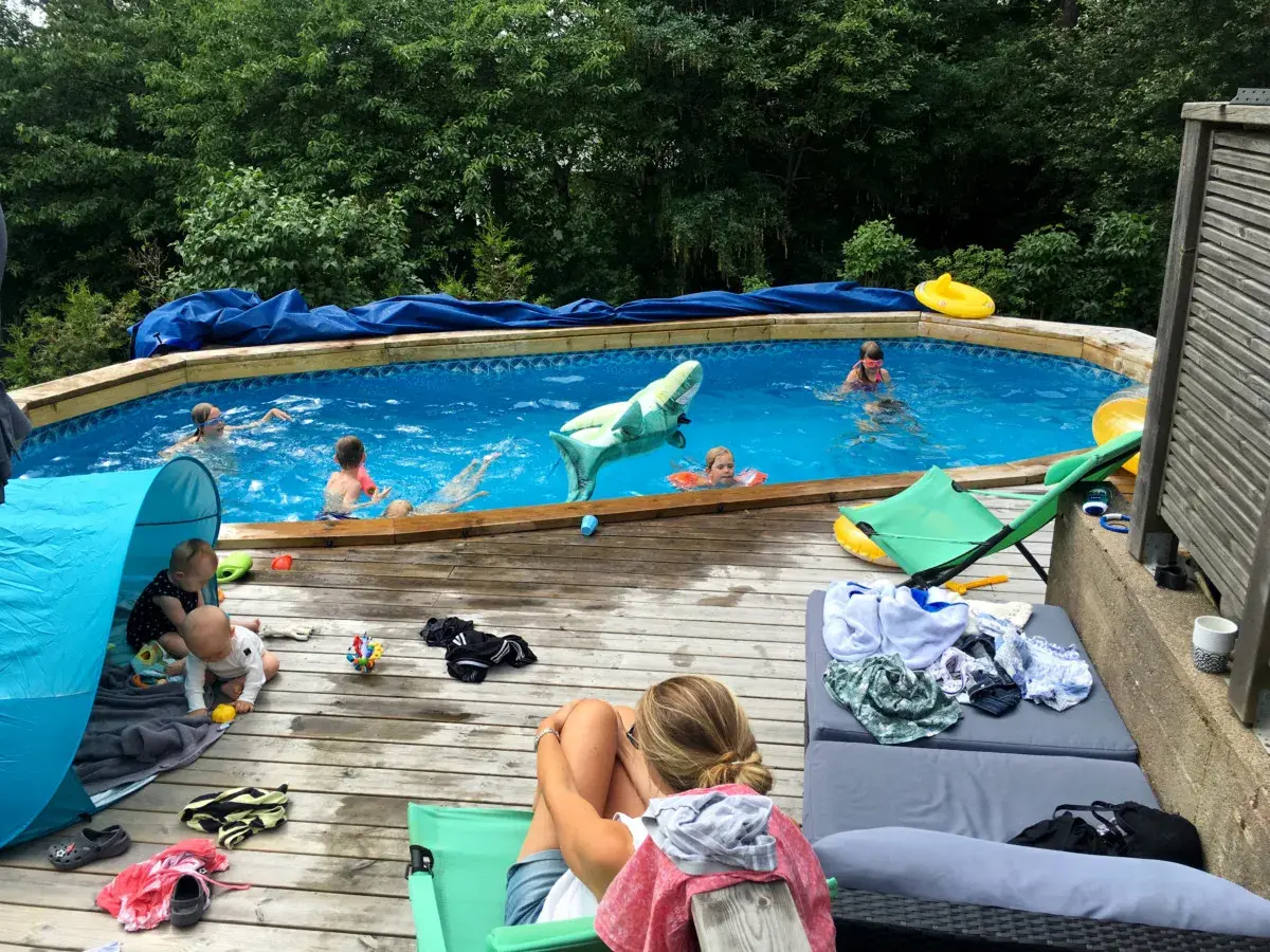 Barn njuter av en solig dag vid poolen, med en uppblåsbar haj och flytleksaker. En bebis leker i ett tält på altanen, perfekt för spf barn.