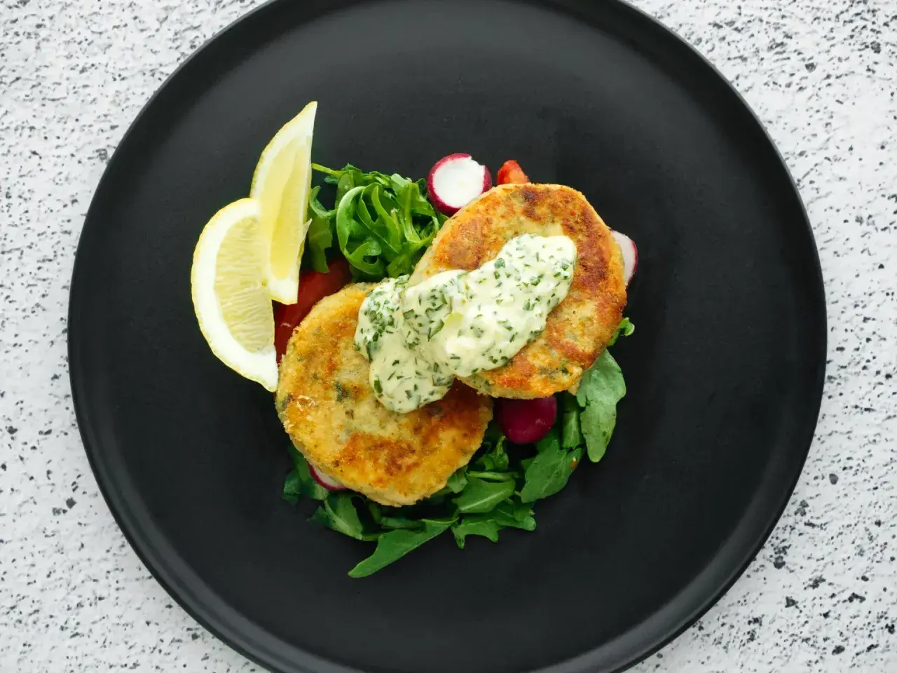 Golden chikuwa tempura patties served with a creamy herb sauce, fresh greens, radishes, and lemon wedges on a black plate.