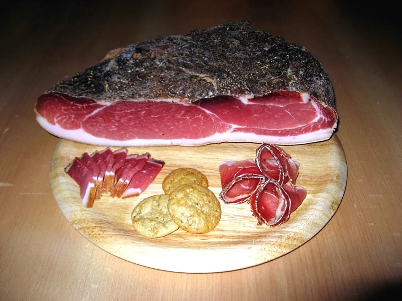 Ein Stück italienischer Speck mit Scheiben und Keksen auf einem Holzbrett.