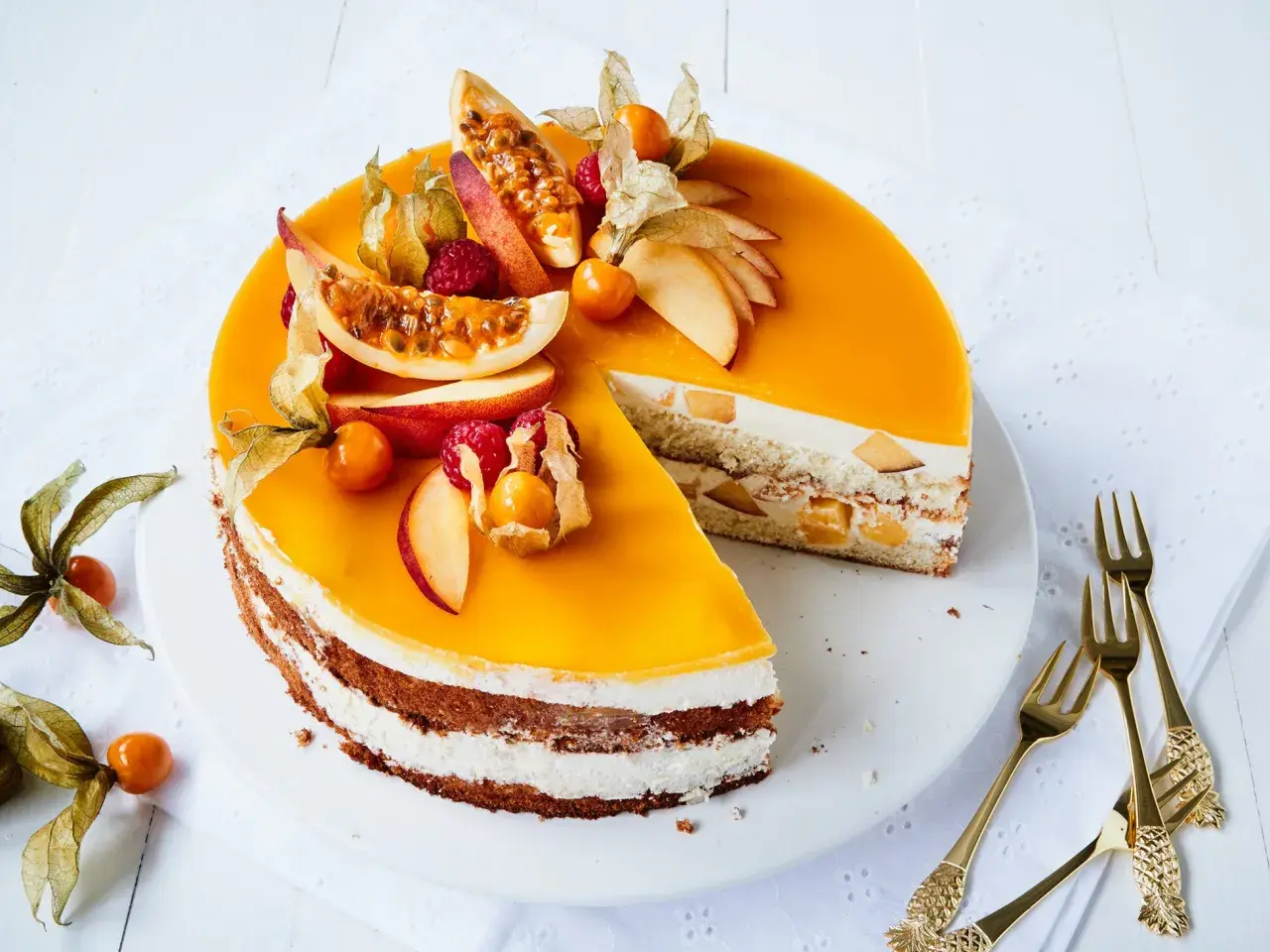 Eine leuchtend gelbe Pfirsich Maracuja Torte, dekoriert mit frischen Früchten und Physalis. Ein Stück ist herausgeschnitten, um die cremige Füllung zu zeigen.