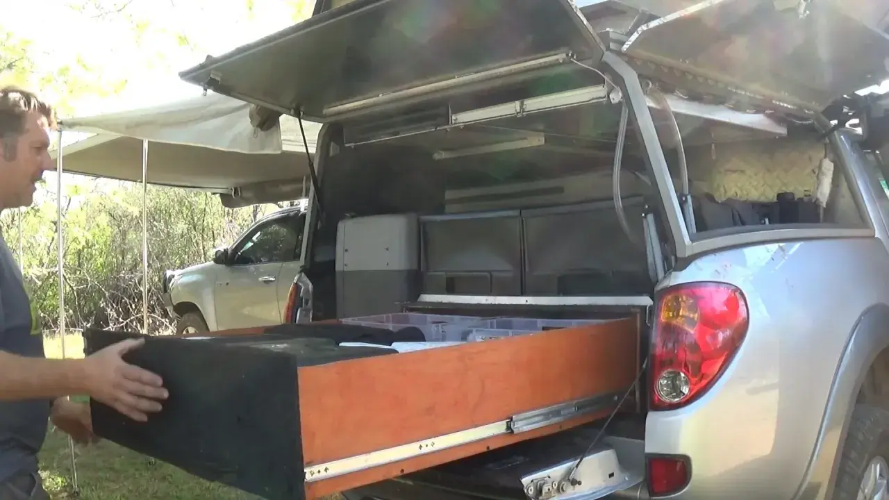 Un homme montre l'intérieur d'une cellule pick-up aménagée avec des tiroirs et des rangements, prête pour l'aventure.