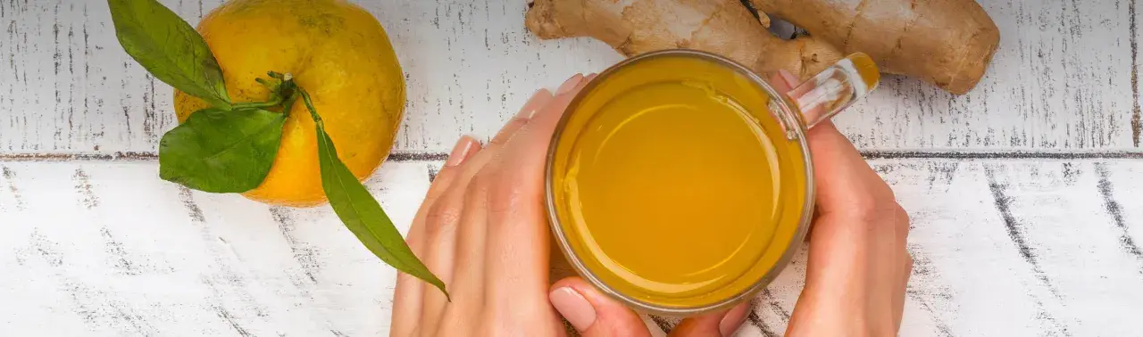 Hände halten ein Glas mit leuchtend gelbem Getränk. Daneben liegen eine Orange und Ingwer. Perfekt, um Kurkuma zuzubereiten.