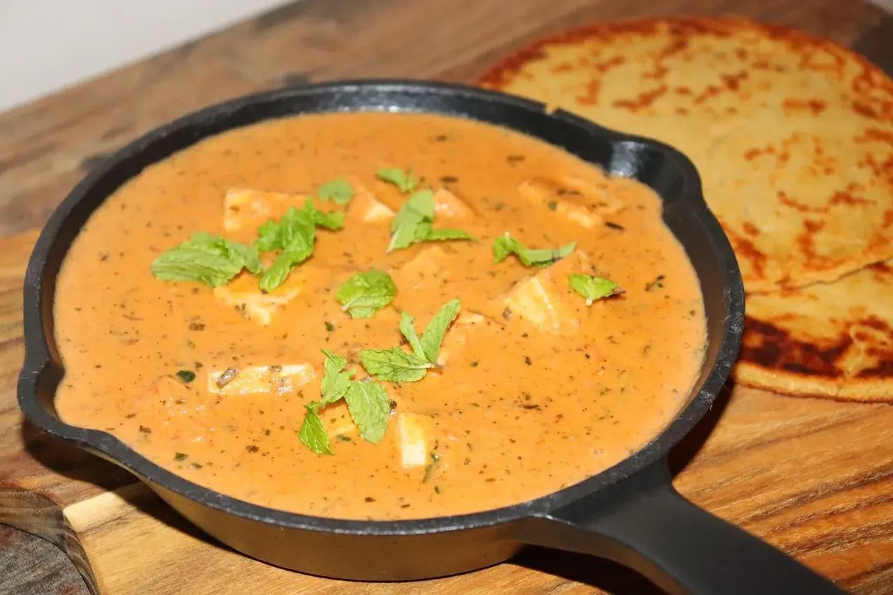 En krämig paneer-rätt i en gjutjärnspanna, garnerad med mynta. Bredvid ligger två gyllene, tunna bröd. Ett perfekt paneer recept!