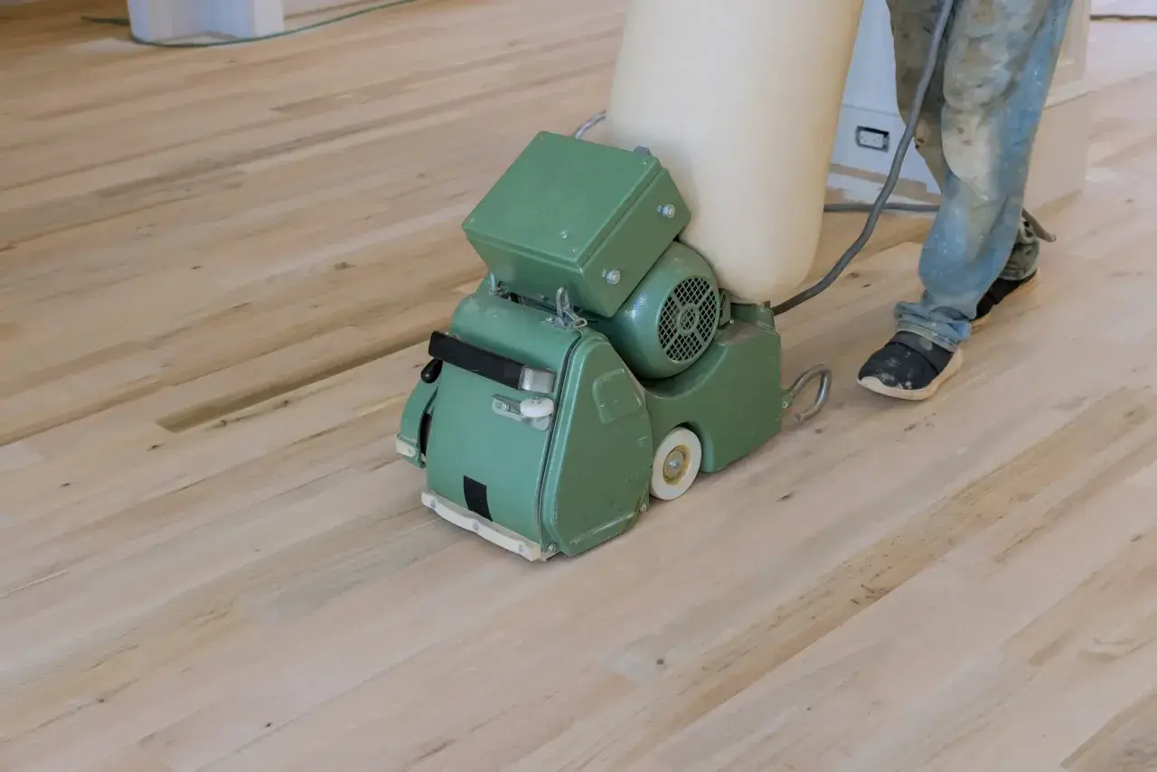 Ein Handwerker schleift mit einer Maschine einen hellen Dielenboden. Staub wird in einem Beutel gesammelt.