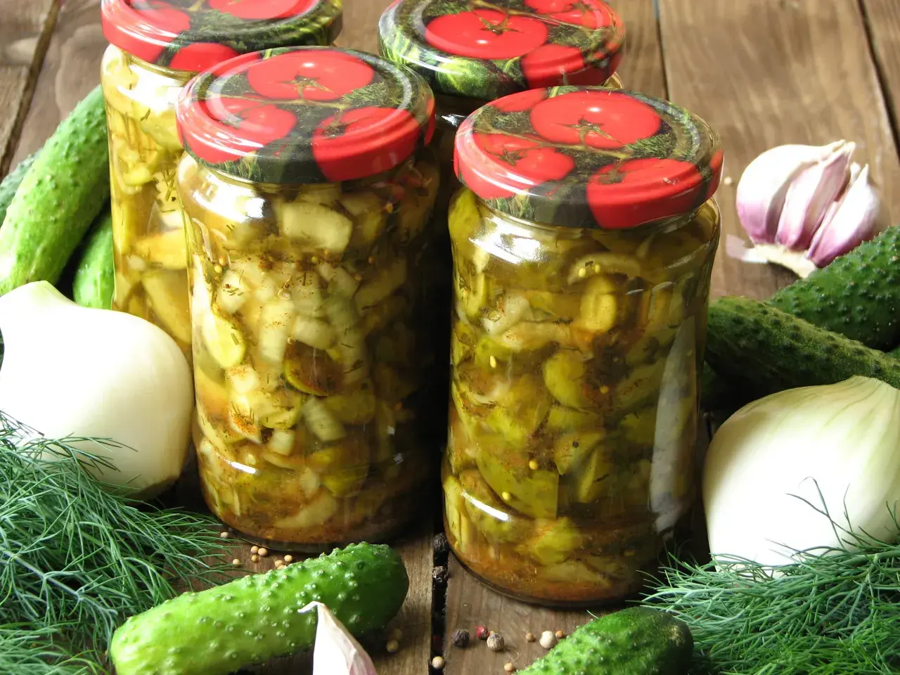 Słoiki z ogórkami w curry ze skórką, otoczone świeżymi ogórkami, cebulą, czosnkiem i koprem.