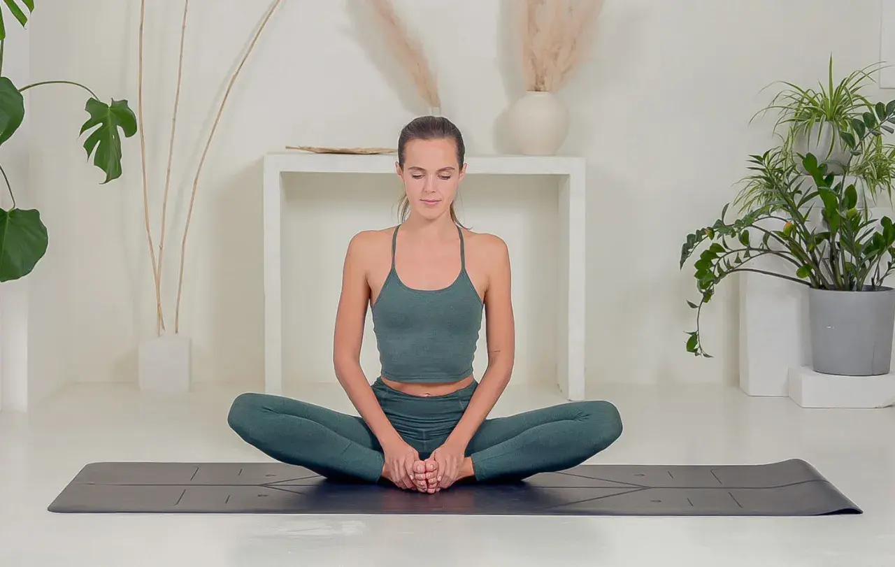 Donna in posizione yoga gambe incrociate, con le mani unite sui piedi, in un ambiente sereno con piante.