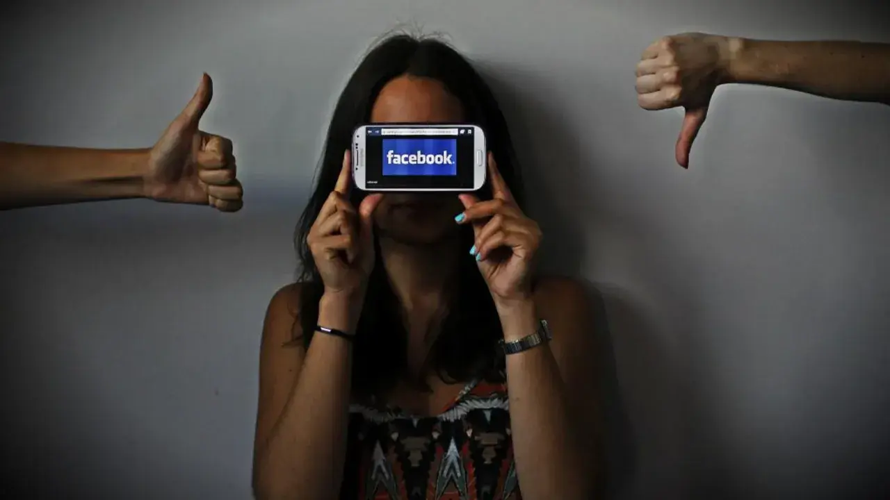 Una persona oculta su rostro tras un móvil con Facebook. A los lados, pulgares arriba y abajo, reflejando el impacto de las redes sociales y la autoestima.
