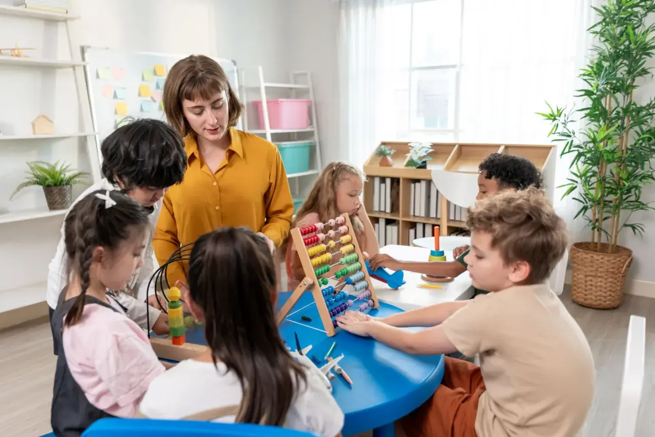 Nauczycielka podczas kursu opiekunki dziecięcej w przedszkolu pomaga dzieciom w zabawie z liczydłem i innymi zabawkami edukacyjnymi.