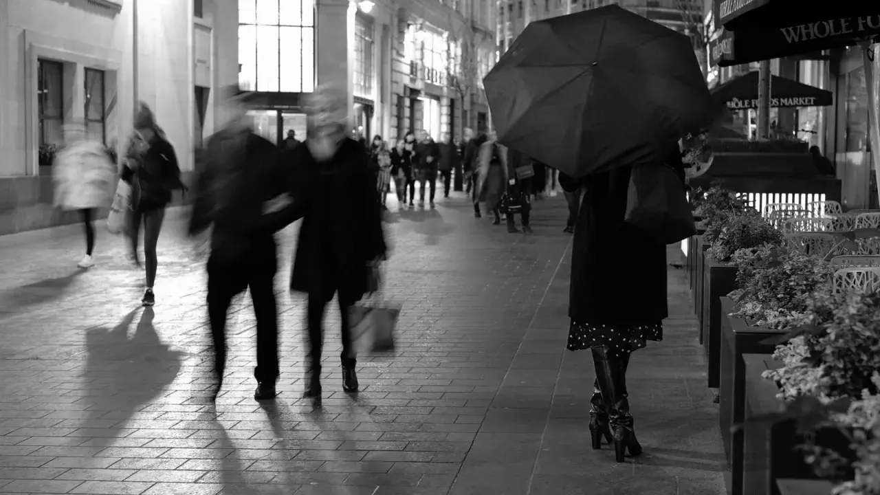 Nocna street photo. Rozmyte postacie spieszą się pod parasolem. W tle widać sklepy i światła miasta.