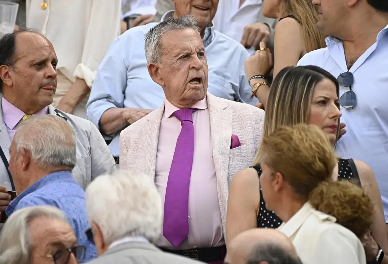Un grupo de personas, entre ellos algunos famosos en las ventas, observan atentamente un evento. El hombre del centro, con traje rosa y corbata morada, parece sorprendido.