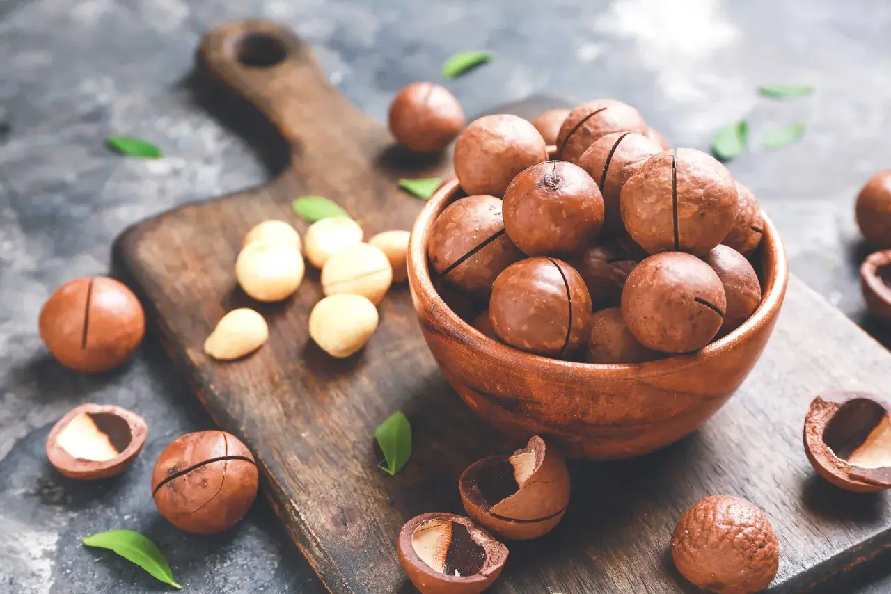 Noci macadamia in una ciotola di legno, con alcune sparse sul tagliere. Scopri i noci macadamia benefici per la tua salute.